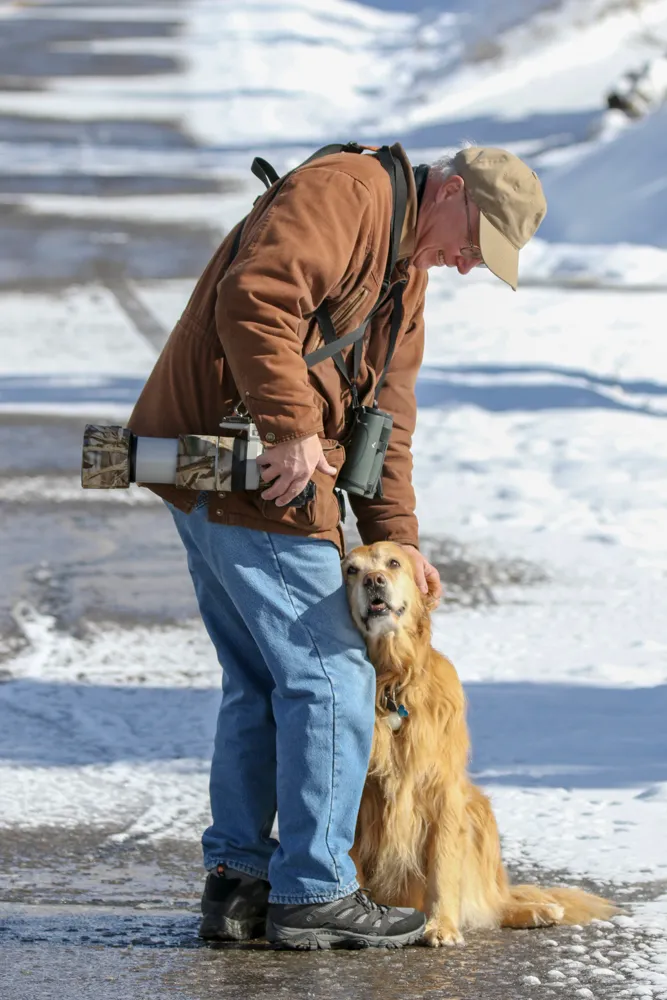 Greg Bodker outdoors with his camera while birding, pausing to greet a dog.